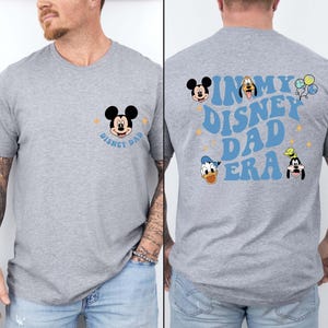 Puede incluir: Una camiseta gris con un gr&aacute;fico de Mickey Mouse en la parte delantera y el texto "In My Disney Dad Era" con gr&aacute;ficos de Mickey Mouse, Donald Duck y Goofy en la parte posterior.