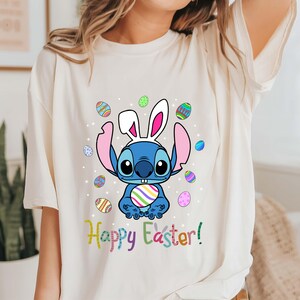 May include: A white t-shirt with a blue Stitch character wearing bunny ears and holding a colorful Easter egg. The text "Happy Easter!" is printed in rainbow colors below the character.