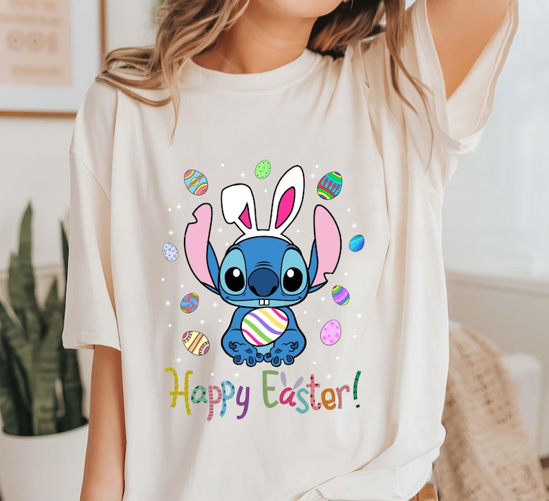May include: A white t-shirt with a blue Stitch character wearing bunny ears and holding a colorful Easter egg. The text "Happy Easter!" is printed in rainbow colors below the character.