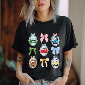 May include: Black t-shirt featuring cartoon car characters in oval frames with colorful bows. The cars are in various colors, including green, red, and blue. The bows are pink, green, yellow, and blue. The shirt is a comfortable, casual style.