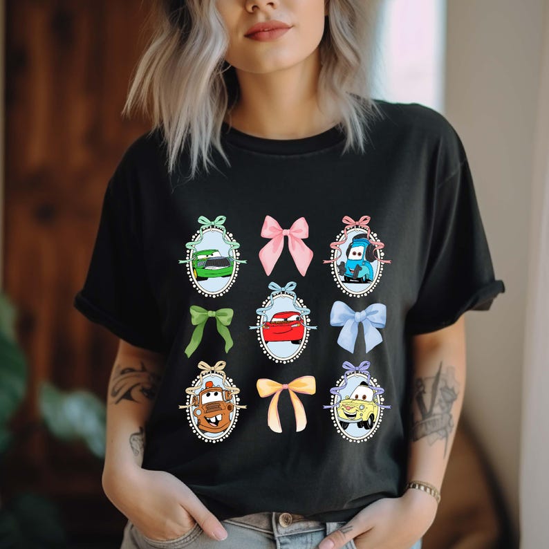 May include: Black t-shirt featuring cartoon car characters in oval frames with colorful bows. The cars are in various colors, including green, red, and blue. The bows are pink, green, yellow, and blue. The shirt is a comfortable, casual style.