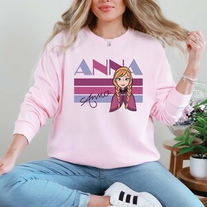 May include: A pale pink sweatshirt with the name "Anna" in purple block letters and a cartoon illustration of a character with blonde braids. The name "Anna" is also written in cursive. The sweatshirt has long sleeves.