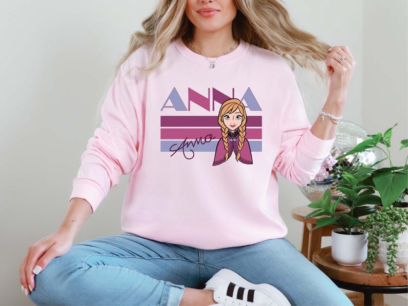 May include: A pale pink sweatshirt with the name "Anna" in purple block letters and a cartoon illustration of a character with blonde braids. The name "Anna" is also written in cursive. The sweatshirt has long sleeves.