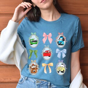 May include: A blue t-shirt featuring cartoon car characters in oval frames with bows. The cars are in various colors, including green, red, and yellow. The bows are pink, green, and blue.