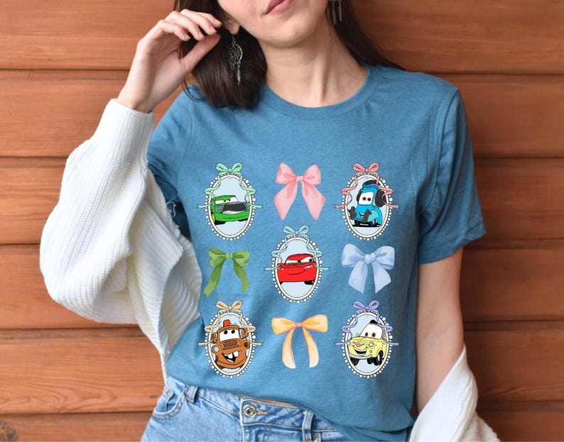 May include: A blue t-shirt featuring cartoon car characters in oval frames with bows. The cars are in various colors, including green, red, and yellow. The bows are pink, green, and blue.