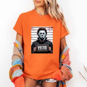May include: Orange t-shirt featuring a graphic of a character in a white mask holding a sign with the number "1978". The character is set against a backdrop of vertical lines, reminiscent of a police lineup. The shirt is short-sleeved.