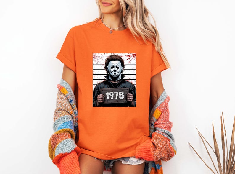 May include: Orange t-shirt featuring a graphic of a character in a white mask holding a sign with the number "1978". The character is set against a backdrop of vertical lines, reminiscent of a police lineup. The shirt is short-sleeved.
