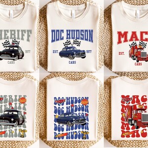 May include: Cream-colored t-shirts featuring cartoon car designs. The shirts display the names "SHERIFF", "DOC HUDSON", and "MACK" in various fonts, along with the text "EST. 1977" and "CARS". Each design includes a car graphic.