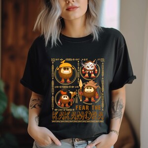 May include: Black t-shirt featuring a graphic design with four cartoon characters and the text "FEAR THE KAKAMORA" in a brown font. The design includes tribal-style borders and details. The shirt is made of a soft material.