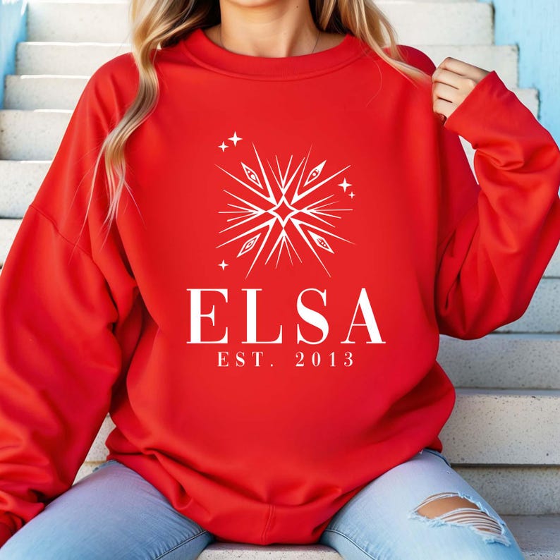 Puede incluir: Sudadera roja con un dise&ntilde;o de copo de nieve blanco y el texto "ELSA EST. 2013"