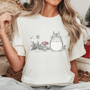 May include: Cream-colored t-shirt featuring a graphic of a large, gray Totoro character with a girl holding a red umbrella at a bus stop. The design is in a minimalist, cartoon style, with black and gray details. The shirt has short sleeves.