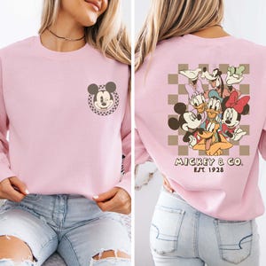 May include: Pink long-sleeved sweatshirt with a Mickey Mouse graphic on the front and a group of Disney characters on the back. The back graphic includes Mickey Mouse, Minnie Mouse, Donald Duck, Goofy, Pluto, and a checkered pattern. The text "Mickey & Co. Est. 1928" is printed below the graphic.