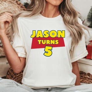 May include: A white short-sleeved t-shirt featuring a graphic print with the text JASON in yellow, TURNS in yellow on a red rectangular background, and the number 5 in yellow with a blue outline, centered on the chest of the garment.