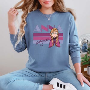 May include: A blue long-sleeved sweatshirt with a graphic of the character Anna from the film Frozen. The design features the name "ANNA" in purple and pink, with Anna's image and signature below.