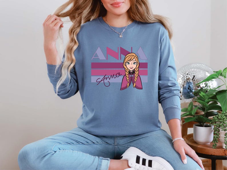 May include: A blue long-sleeved sweatshirt with a graphic of the character Anna from the film Frozen. The design features the name "ANNA" in purple and pink, with Anna's image and signature below.