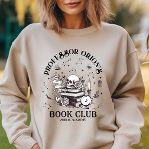 May include: Beige sweatshirt with a black and white graphic. The graphic features a skull, books, roses, a candle, and the text "Professor Orion's Book Club Zodiac Academy."