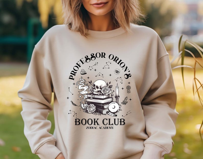 May include: Beige sweatshirt with a black and white graphic. The graphic features a skull, books, roses, a candle, and the text "Professor Orion's Book Club Zodiac Academy."