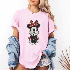 May include: A pink t-shirt with a cartoon image of Minnie Mouse wearing a red bow with white polka dots. The image is in a vintage style.