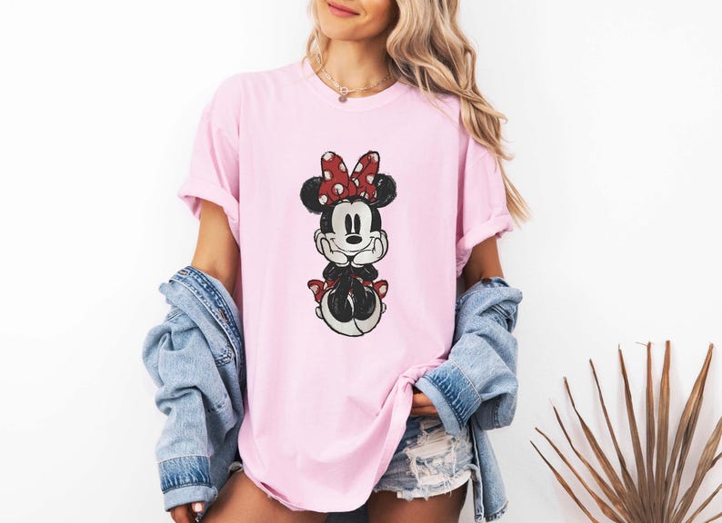 May include: A pink t-shirt with a cartoon image of Minnie Mouse wearing a red bow with white polka dots. The image is in a vintage style.