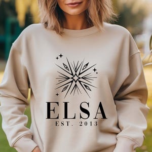 Puede incluir: Una sudadera beige con un dise&ntilde;o gr&aacute;fico negro que presenta una estrella estilizada con ojos y el texto "ELSA EST. 2013".