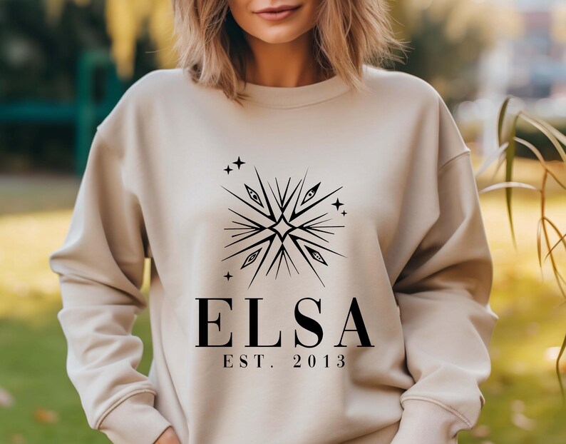 Puede incluir: Una sudadera beige con un dise&ntilde;o gr&aacute;fico negro que presenta una estrella estilizada con ojos y el texto "ELSA EST. 2013".