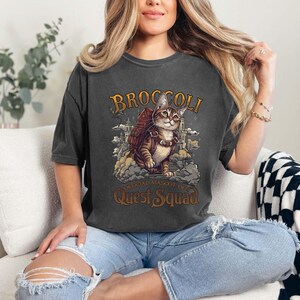 Pu&ograve; includere: Una maglietta grigia scura con una grafica in stile vintage di un gatto con uno zaino e il testo "Broccoli, mascotte ufficiale del Quest Squad".