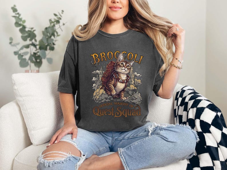Pu&ograve; includere: Una maglietta grigia scura con una grafica in stile vintage di un gatto con uno zaino e il testo "Broccoli, mascotte ufficiale del Quest Squad".