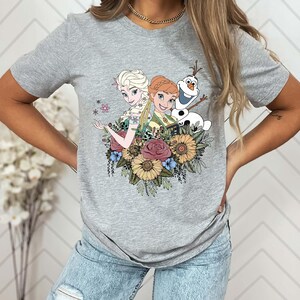 May include: Gray t-shirt with a graphic print of Elsa and Anna from Frozen, along with Olaf, surrounded by flowers.