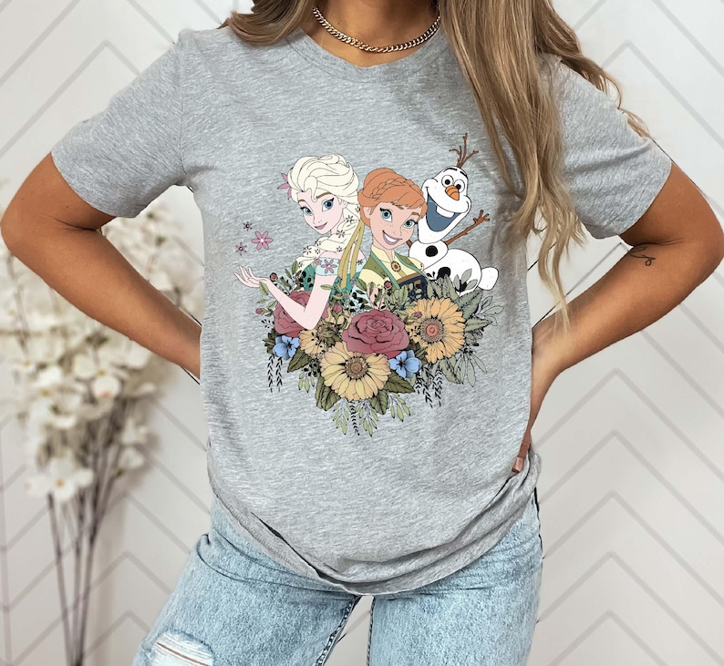 May include: Gray t-shirt with a graphic print of Elsa and Anna from Frozen, along with Olaf, surrounded by flowers.