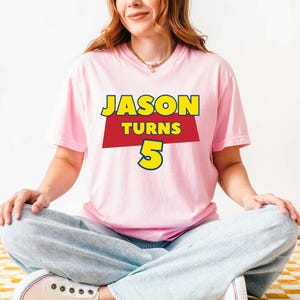 May include: A light pink short-sleeved t-shirt featuring a graphic design with the yellow text JASON, the red text TURNS, and the large yellow number 5. The shirt is paired with light blue denim jeans and white sneakers.