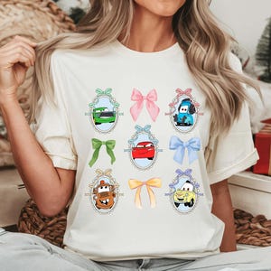 May include: Cream-colored t-shirt featuring a design of cartoon cars in oval frames with bows. The cars are in various colors, including green, red, blue, and yellow. The bows are in shades of pink, green, and yellow.