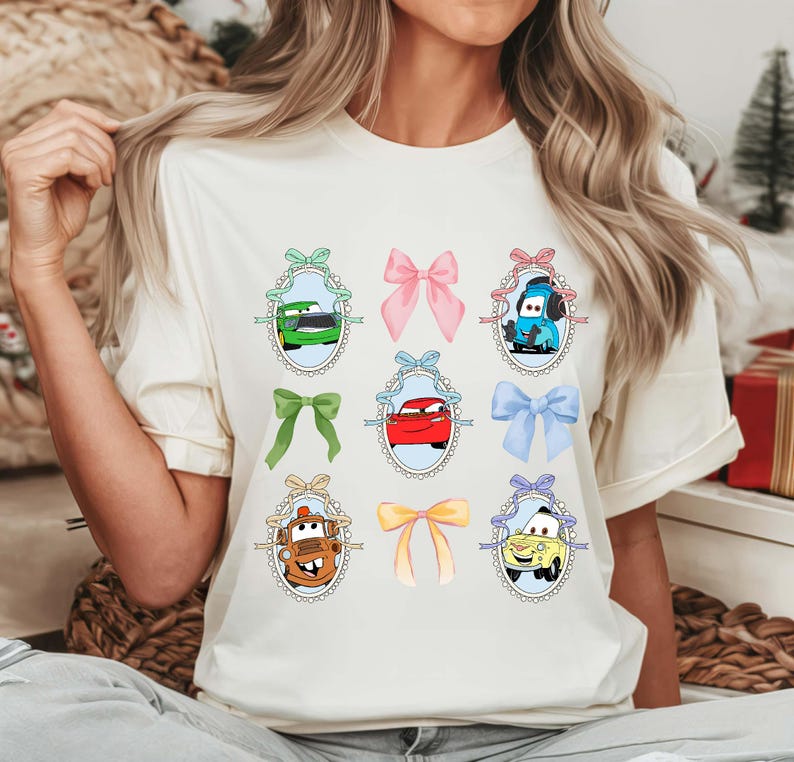 May include: Cream-colored t-shirt featuring a design of cartoon cars in oval frames with bows. The cars are in various colors, including green, red, blue, and yellow. The bows are in shades of pink, green, and yellow.
