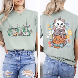 May include: A light green t-shirt with a graphic print of three cats in front of the Eiffel Tower. The cats are wearing a bow tie, a collar, and a bow. The t-shirt has a crew neck and short sleeves.