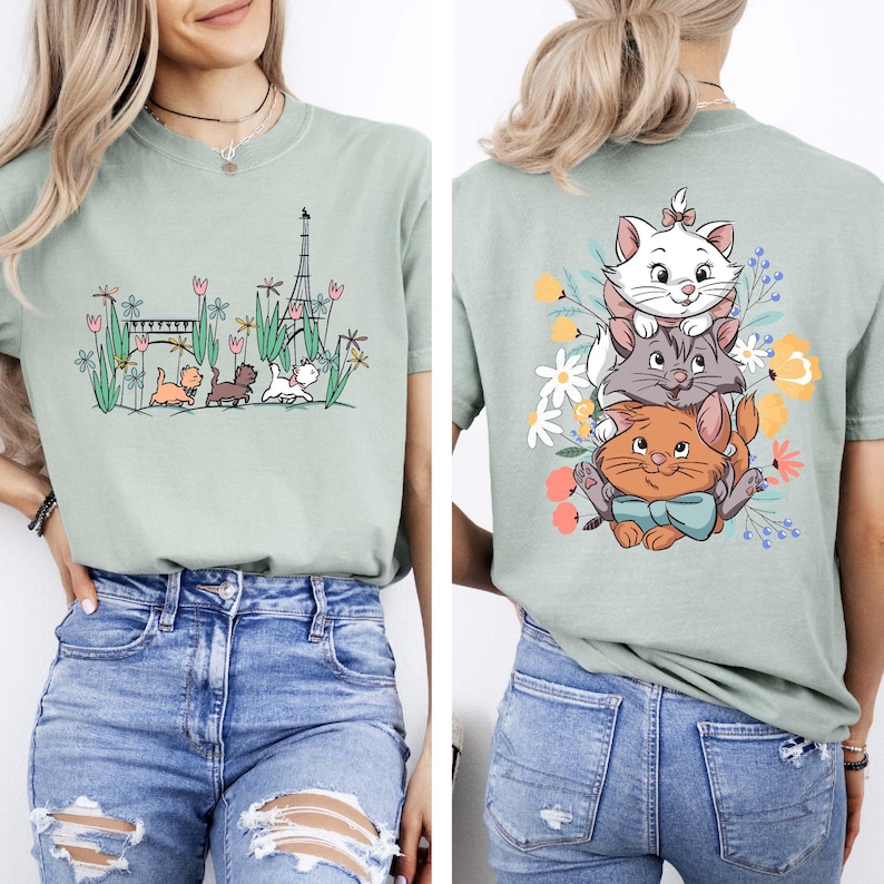 May include: A light green t-shirt with a graphic print of three cats in front of the Eiffel Tower. The cats are wearing a bow tie, a collar, and a bow. The t-shirt has a crew neck and short sleeves.
