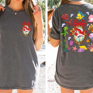 May include: A gray t-shirt with a cartoon image of Ariel, the Little Mermaid, in a frame surrounded by seashells, starfish, and other ocean-themed imagery.