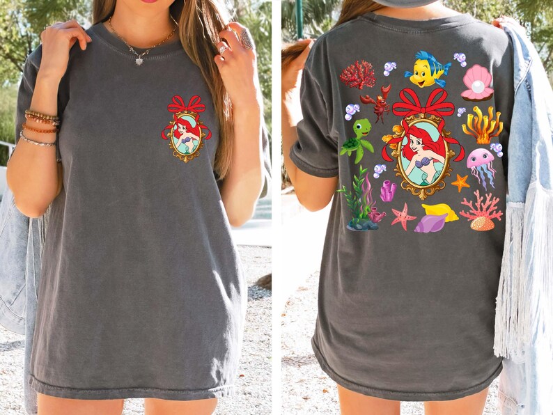 May include: A gray t-shirt with a cartoon image of Ariel, the Little Mermaid, in a frame surrounded by seashells, starfish, and other ocean-themed imagery.