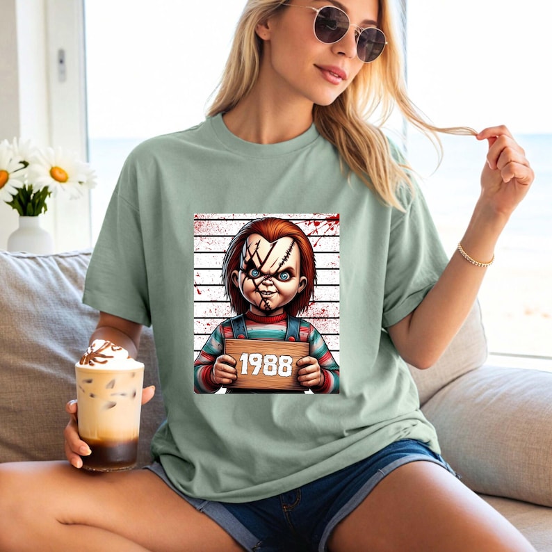 May include: Sage green t-shirt featuring a graphic of a doll with stitched scars and the year "1988" on a wooden sign. The doll has red hair, blue eyes, and is wearing striped overalls. The background includes a blood spatter design.
