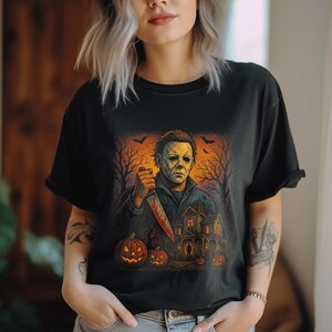 May include: Black t-shirt featuring a Halloween-themed graphic. The design includes a menacing figure holding a bloody knife, a haunted house, jack-o'-lanterns, and bare trees against an orange and black background. Bats are flying in the sky.