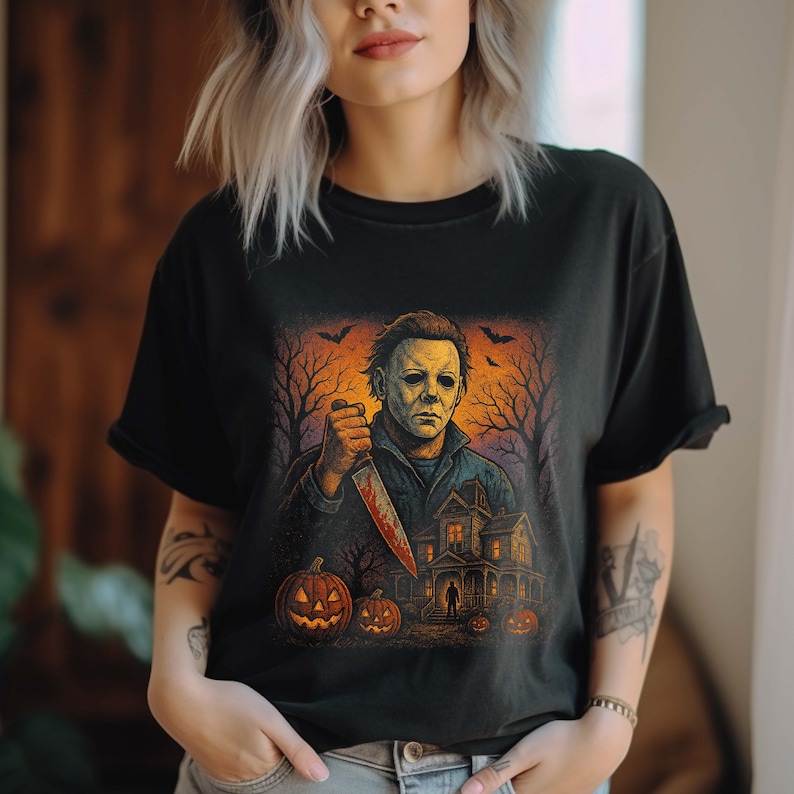 May include: Black t-shirt featuring a Halloween-themed graphic. The design includes a menacing figure holding a bloody knife, a haunted house, jack-o'-lanterns, and bare trees against an orange and black background. Bats are flying in the sky.