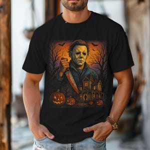 May include: Black t-shirt featuring a Halloween-themed graphic. The design includes a masked figure holding a bloody knife, a haunted house, pumpkins, and bats against an orange and purple sky. The artwork has a vintage, horror movie aesthetic.