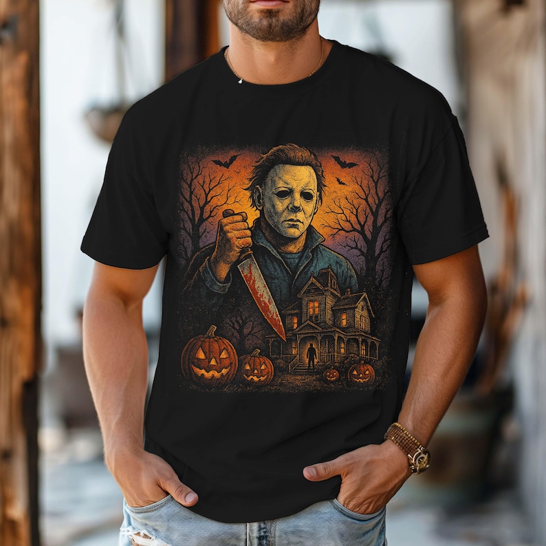 May include: Black t-shirt featuring a Halloween-themed graphic. The design includes a masked figure holding a bloody knife, a haunted house, pumpkins, and bats against an orange and purple sky. The artwork has a vintage, horror movie aesthetic.
