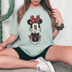 May include: A light blue t-shirt with a vintage-style image of Minnie Mouse wearing a red bow. The shirt is being worn by a person sitting on a white surface.