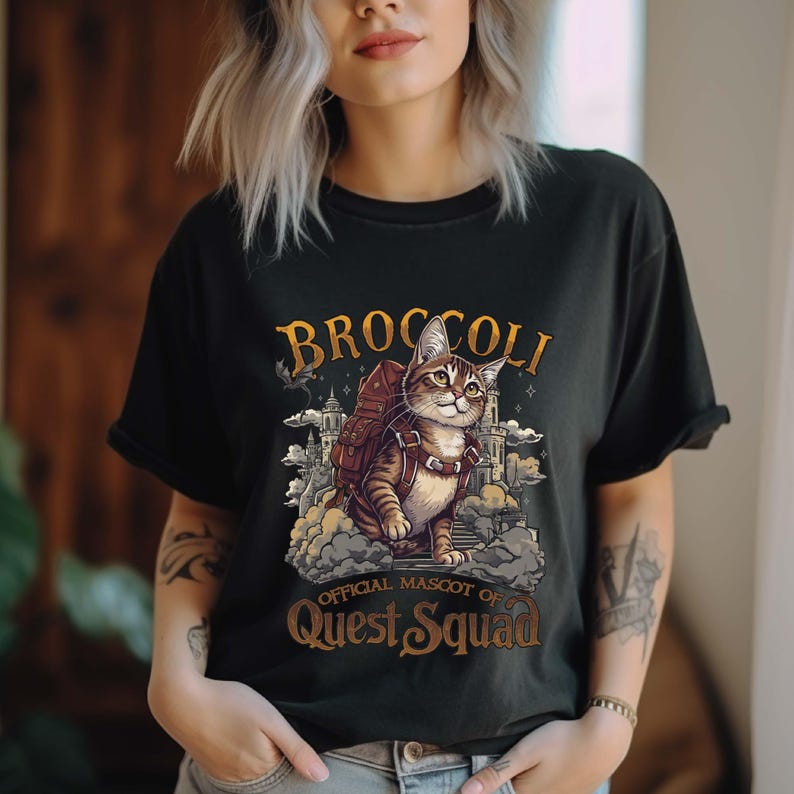 Pu&ograve; includere: Maglietta nera con una grafica di un gatto che indossa uno zaino e sta in piedi su una nuvola. Il testo "Broccoli, mascotte ufficiale di Quest Squad" &egrave; stampato sulla maglietta.
