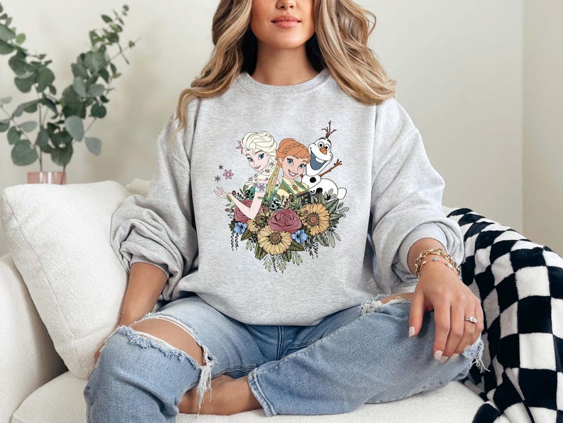Pu&ograve; includere: Una felpa grigia con una stampa grafica di Elsa e Anna di Frozen, insieme a Olaf, circondato da fiori.