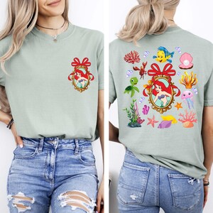 May include: Two light green t-shirts with a cartoon image of Ariel from The Little Mermaid. One shirt has a single image of Ariel in a frame with a red bow. The other shirt has multiple images of Ariel, sea creatures, and seashells.