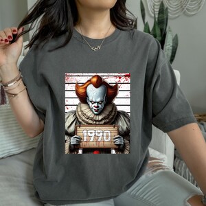 May include: Dark gray t-shirt featuring a graphic of a scary clown holding a sign that reads "1990". The clown has white face paint, red hair, and a ruffled collar. The background includes a police lineup and blood splatter.