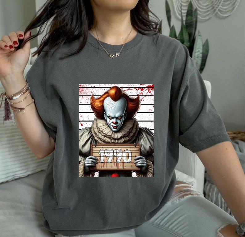 May include: Dark gray t-shirt featuring a graphic of a scary clown holding a sign that reads "1990". The clown has white face paint, red hair, and a ruffled collar. The background includes a police lineup and blood splatter.