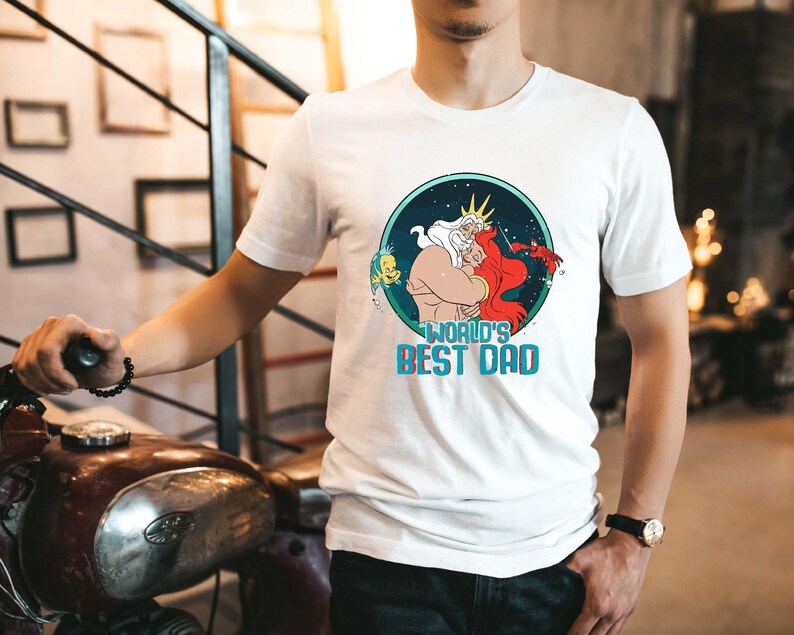 May include: White t-shirt with a graphic of King Triton from The Little Mermaid hugging Ariel. The graphic is in a circle with a blue background and stars. The text "World's Best Dad" is below the graphic.