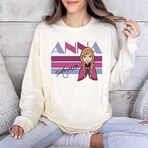 May include: Cream-coloured long-sleeve sweatshirt with the name "ANNA" in purple and blue block letters above a cartoon illustration of a character with blonde braids. The name "Anna" is also written in cursive below the illustration.