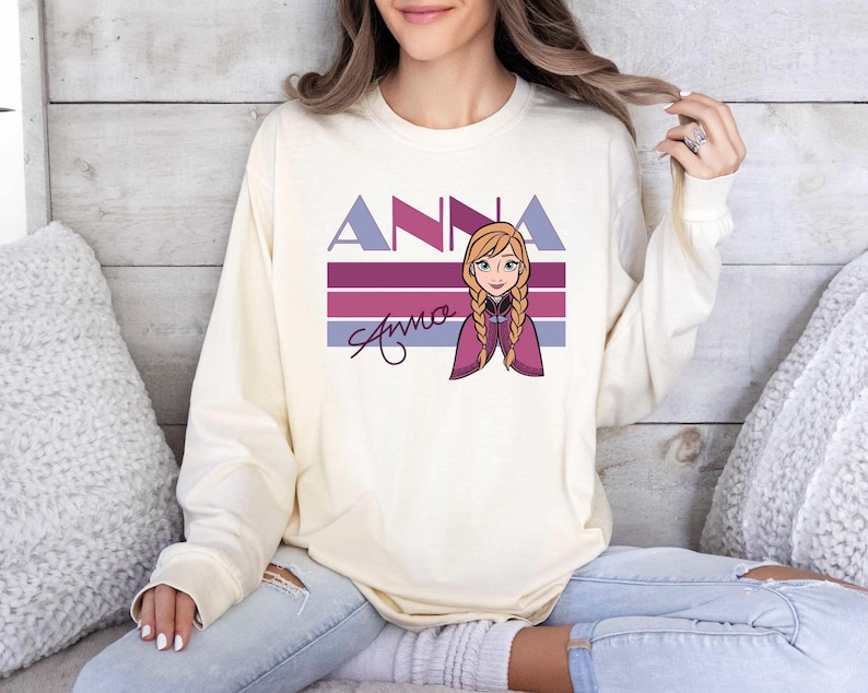 May include: Cream-coloured long-sleeve sweatshirt with the name "ANNA" in purple and blue block letters above a cartoon illustration of a character with blonde braids. The name "Anna" is also written in cursive below the illustration.